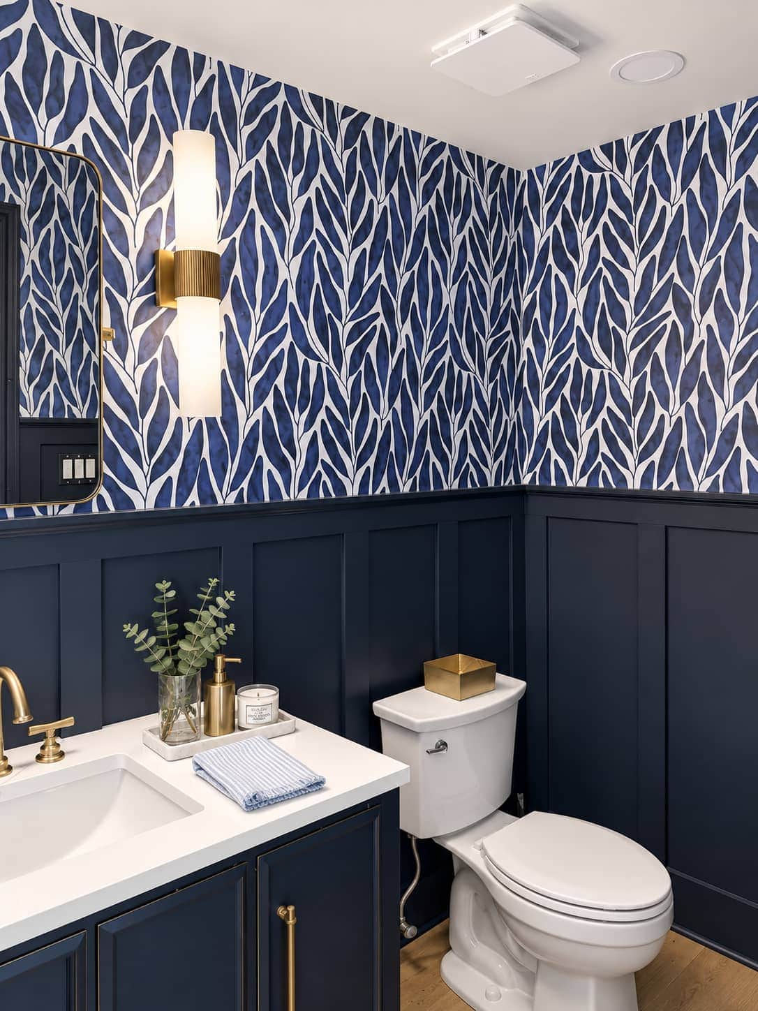 Powder room with navy board-and-batten wainscoting, blue leaf wallpaper, a brass sconce and a white quartz vanity