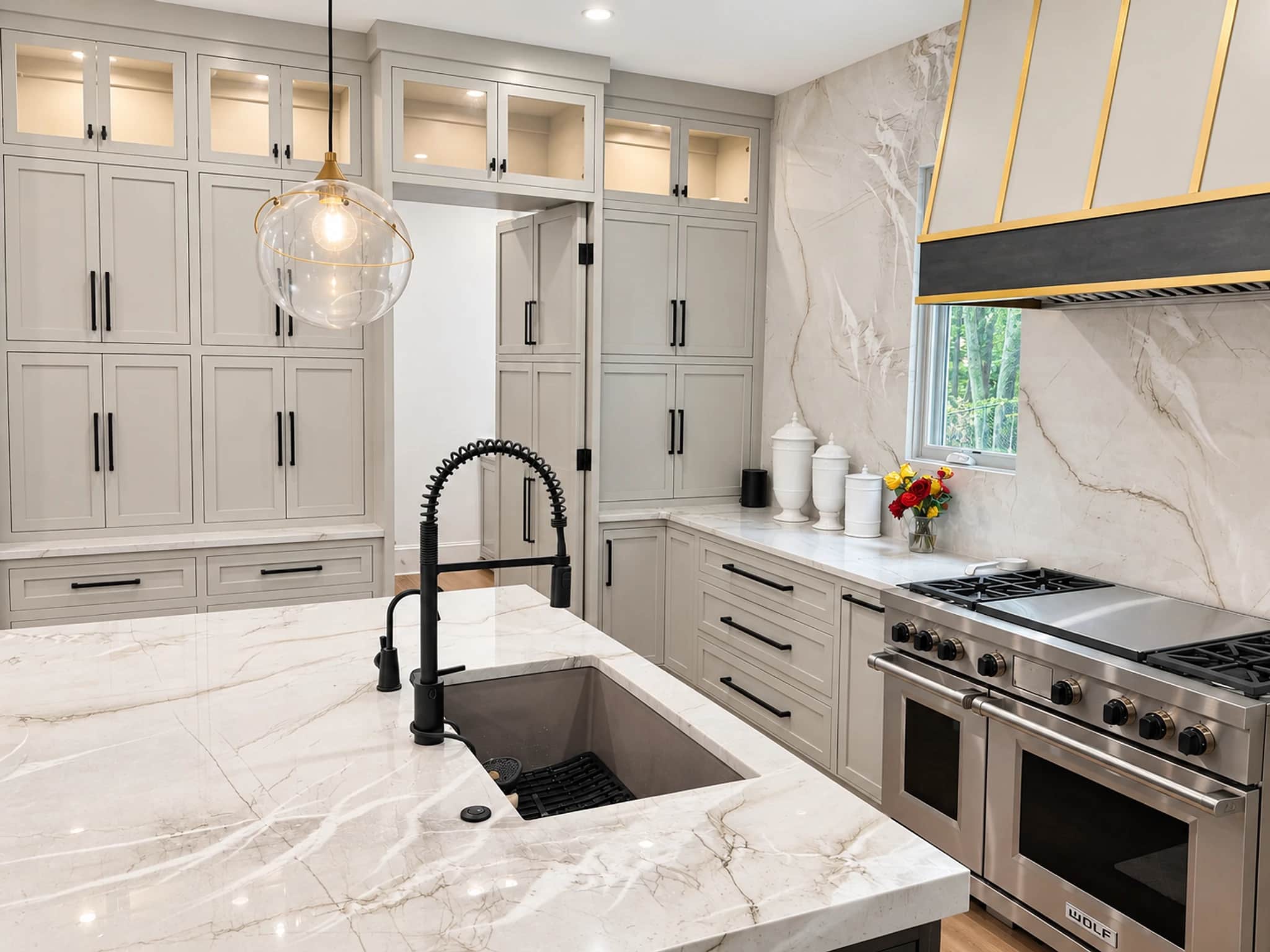 Remodeled kitchen with greige inset cabinetry, a marble-topped island, lit glass-front upper cabinets and a Wolf range under a brass-banded hood