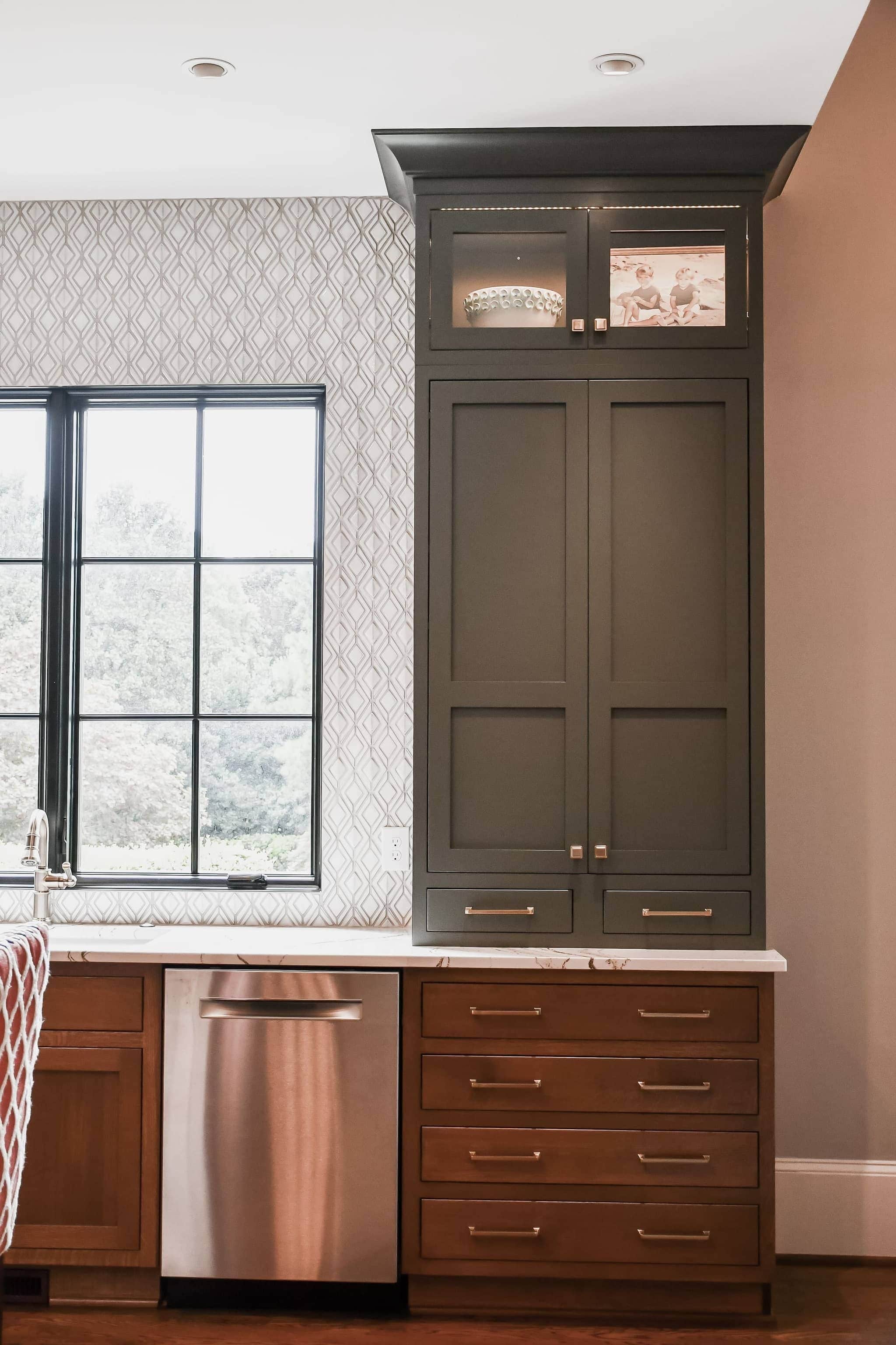 Kitchen cabinetry in olive and cherry with lit glass-front uppers, brass pulls and a diamond-pattern tile backsplash