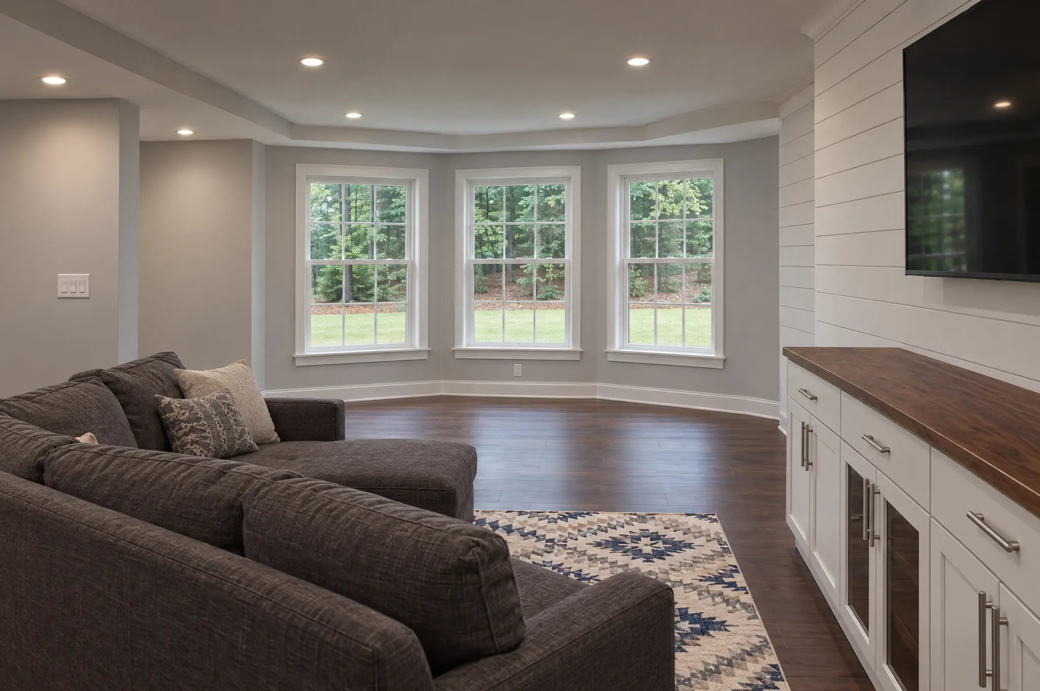 Finished daylight basement with a bay of three double-hung windows facing a wooded yard, a gray sectional, and a white shiplap wall with a mounted TV over a shaker media cabinet with a walnut top