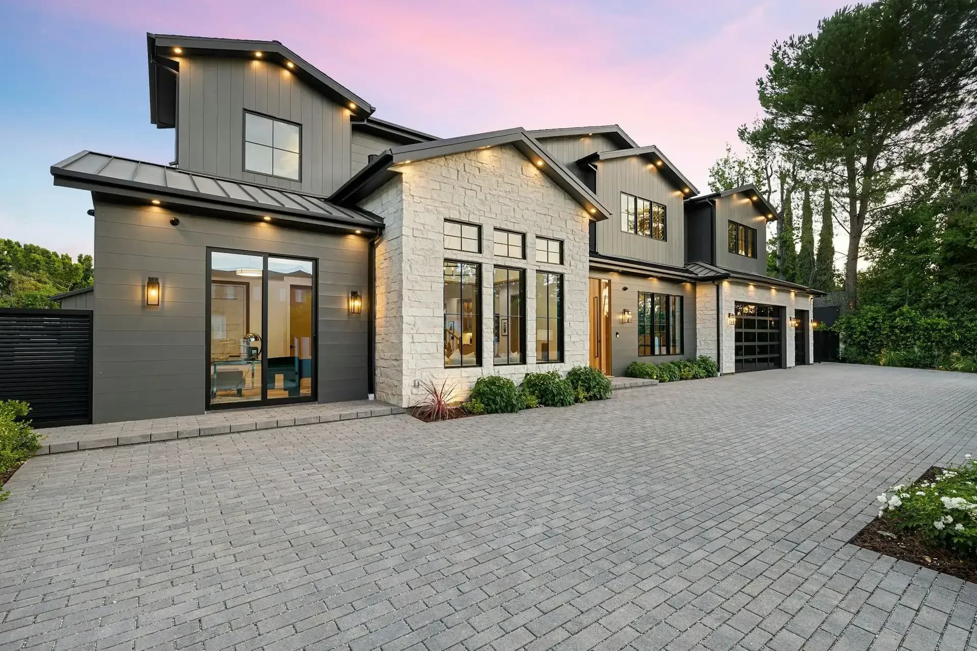 Modern farmhouse exterior at dusk with gray siding, white stone, black windows and a paver drive