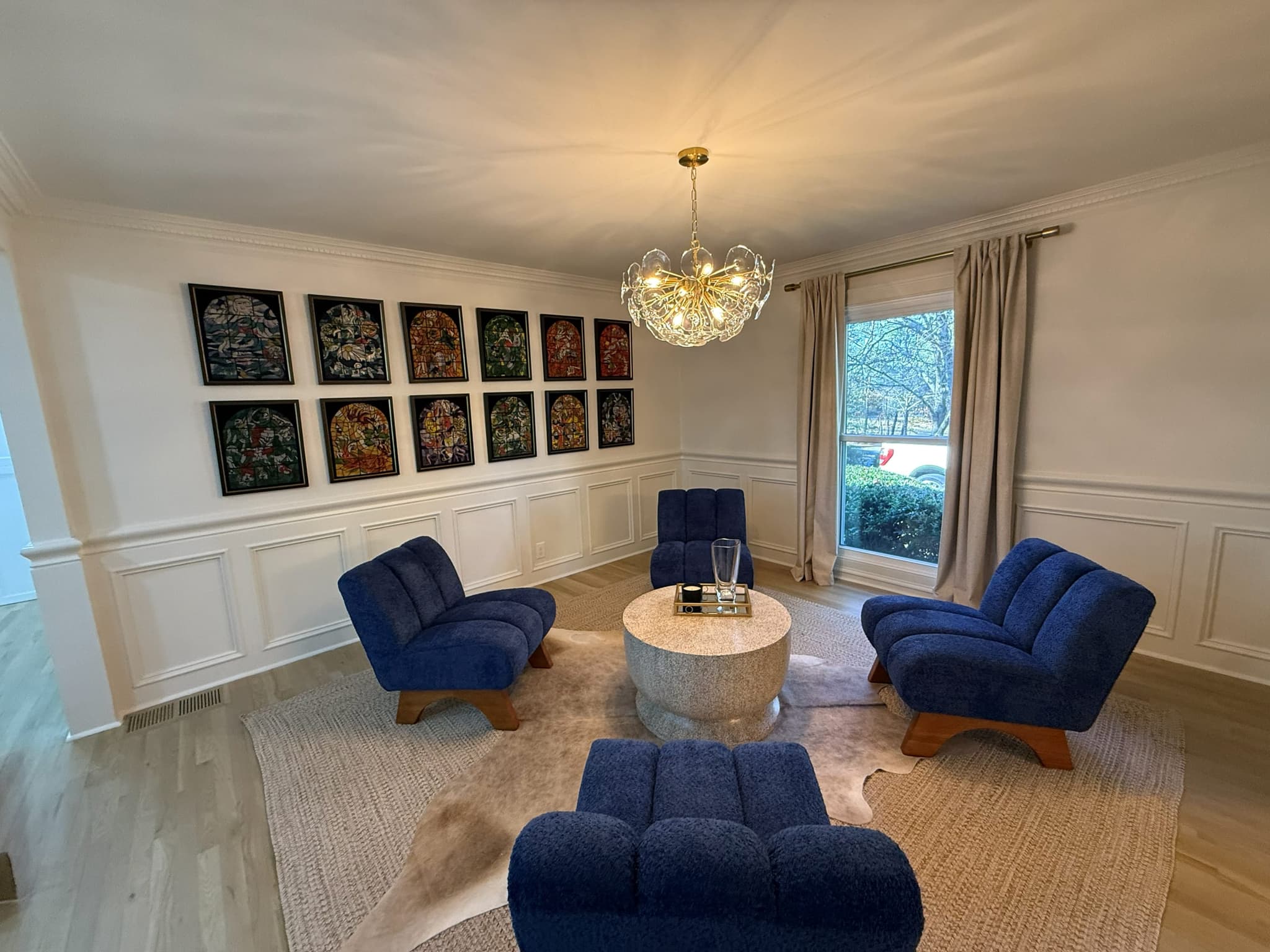 Formal sitting room with picture-frame wainscoting, a gallery wall of framed stained-glass prints and blue lounge chairs around a round stone table
